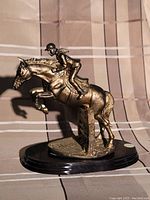 Bronze-colored sculpture showing a jockey riding a horse, mid-jump over a hurdle, featuring detailed musculature and attire, mounted on a black polished oval base.