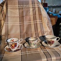Three tea cup and saucer sets with varied floral and fruit patterns on beige background.