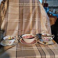 Front view of three Aynsley bone china tea cups with matching saucers, each featuring distinct floral patterns and gold accents, displayed on fabric background.