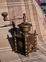 Full view of the vintage brass coffee grinder showing the crank handle with wooden knob, decorative columns, floral side detailing, and drawer front.