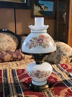 Photo shows a vintage milk glass hurricane lamp with floral patterns, brass base, and a frosted chimney, sitting on a patterned surface in a room with traditional furnishings.