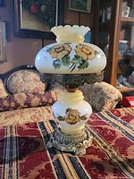 Full view of vintage white glass hurricane style electric lamp with floral painting and decorative metal elements on the base and rim of shade.