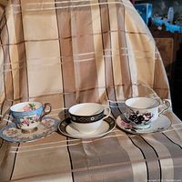 Three tea cups and saucers displayed on checked cloth background showing floral and geometric designs