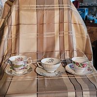 Three bone china tea cups and saucers displayed on a plaid cloth showing different floral designs and gold trims.
