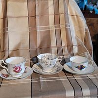 Three different tea cups and saucers displayed together on fabric background, showing individual designs and patterns.