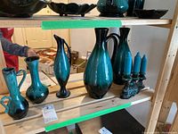 Six Blue Mountain Pottery ceramic pieces including various vases and pitchers displayed on shelf. Glossy teal and black drip glaze coloration.