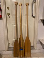 Three wooden boating paddles standing upright against a door, varying in finish and condition.