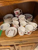 Full view of multiple teacups, saucers, and small plates arranged on wooden furniture shelf showing various floral designs and patterns