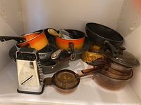 Photo of various cookware items showing amber Vision Ware glass pots, vintage enamelware colored in shades of orange/yellow, black pans, and kitchen utensils inside a white storage shelf.