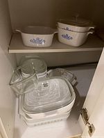White Corning Ware casserole dishes with Blue Cornflower pattern on middle and upper cabinet shelves, stacked with clear glass lids, and several clear glass Glasbake and Fire King dishes below.