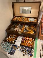 Full view of open wooden jewelry box with multiple sections filled with assorted costume jewelry including earrings, necklaces, and bracelets. Jewelry box lid has a mirror and a label indicating Lot 45.