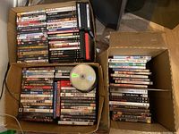 Three cardboard boxes filled with DVDs including loose discs on spindle with covers visible, showing variety of movies in action, comedy, and romance.
