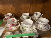 Group photo showing six fine china teacups and matching saucers in floral patterns on a wood shelf.