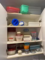 Three shelves inside a white cabinet containing a variety of plastic and glass food storage containers in assorted sizes with red, white, blue, orange, and beige lids.