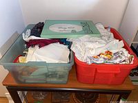 Two storage bins filled with assorted vintage linens including aprons, handkerchiefs, embroidered linens, and other textile items.