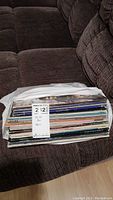 Stack of approximately 24 LP vinyl records in sleeves on a brown couch, with MaxSold tag visible showing quantity and partial description