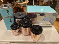Full view of seven retro kitchen containers including turquoise tin canisters, three copper tone canisters with black lids, and matching turquoise bread box with chrome accents.