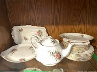 Shelved display of Royal Albert Lily of the Valley china including teapot, serving platters, and stacked plates with floral design and gold trim
