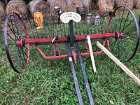 View of antique red and black dump rake with adjustable teeth and white metal operator seat, set on grassy ground with hay bales in background.