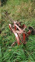Red steel flail mower implement resting in overgrown grass, showing overall shape and mounting frame