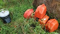 Five orange gasoline cans and one black bucket on grass near hay.