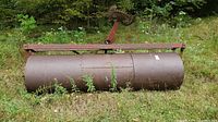 Front view of vintage steel field roller with rust and attached spiked wheel, situated in a field.
