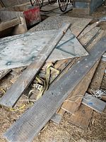 Photo showing assorted long barn boards and plywood sheets stacked on barn floor with hay and dirt visible. Wooden planks have a weathered look with gray and brown patina.