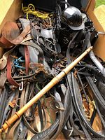 Top view of a box filled with various horse tack including leather harnesses, reins, a wooden crop, and a black bull riding helmet.