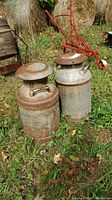 Two vintage metal milk cans outdoors on grass, showing rust and aged patina.