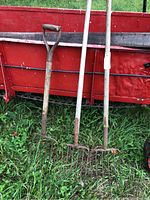 Three vintage pitchforks leaning against farm equipment outdoors on grass.