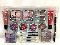 Full set of The Color Workshop makeup cosmetics in sealed clear plastic packaging showing eyeshadows, lip glosses, eye pencil, brushes, and sponges arranged neatly.