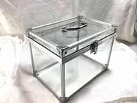 Closed view of the transparent display box showing front latch, handle on lid, metal corner reinforcements, and clear acrylic sides.
