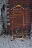 Full view of vintage wooden valet stand against brick wall showing hanger, tray, and metal base with wheels.