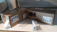 Two sealed cardboard boxes on a shelf, labeled 3M containing the eyewear. One pair of clear framed glasses and one sealed 3M branded plastic bag with eyewear are visible in front of the boxes.