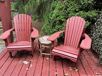 Two red Adirondack chairs with small wooden side table placed outside on a red wood deck surrounded by greenery.
