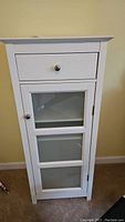 Front view of white wooden cabinet with single drawer and three-pane glass door, showing brushed nickel knobs.
