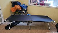 Black camp cot set up on carpet with lounge chair, sleeping bags and mats in background