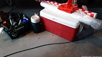 Photo shows red and white Coleman cooler, white picnic table plastic covers rolled and placed on top, and green crate with assorted dark bottles and containers.