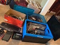 Photo showing assortment of electronic items including a monitor, keyboard, cables in a blue storage bin and a cardboard box.