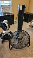 Four fans positioned on carpet in room with window air conditioner. Black cabinet and speaker also in background. Fans vary in style and size including tower and circular fans.