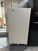 Closed white Frigidaire upright freezer positioned between a wall and a refrigerator in a garage setting.