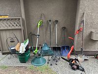 Wide shot showing garden tools lined against wall including rakes, shovels, garden basket, and a green string trimmer.