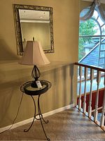 Metal table with glass top, black metal base, table lamp with black metal base and beige fabric shade with visible hole, table and lamp shown together against wall