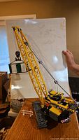 Side view of assembled yellow Lego crawler crane holding a green and white windowed container using pulley and hook system.