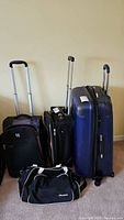 Four luggage pieces including a large blue hardside suitcase, two black softside rolling suitcases, and one black duffel bag arranged on floor near wall.