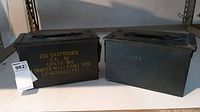 Two green rectangular metal ammunition boxes placed side by side on shelf. One box has yellow stencil text describing 100 linked M9 tracer cartridges.