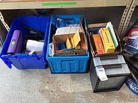 Long shot showing three storage containers including two blue plastic bins, one cardboard box with paper products, and a small black stacking file drawer on the floor.