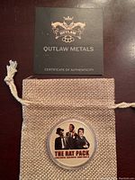 Photo of coin inside clear protective case with The Rat Pack colored artwork visible, placed on tan woven pouch with Outlaw Metals Certificate of Authenticity card above.
