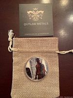 Customized silver coin featuring Deadpool image inside clear protective capsule, laying on burlap pouch, with Outlaw Metals certificate of authenticity on top