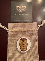 Outlaw Metals certificate of authenticity card above a beige burlap drawstring pouch containing the coin.